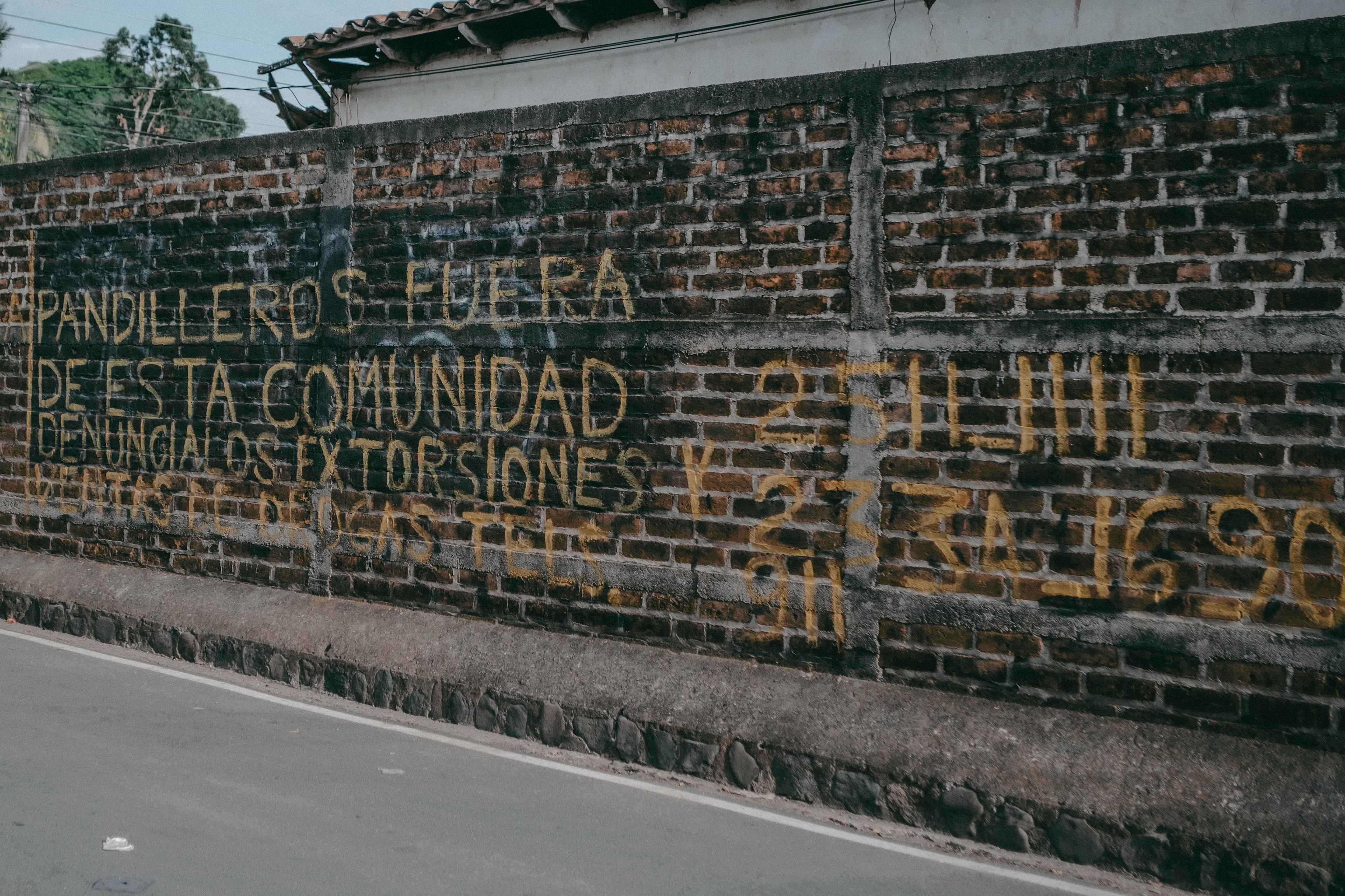 ''Pandilleros fuera de esta comunidad. Denúncialos'', se lee en el muro de una comunidad donde algunos familiares de personas detenidas durante el régimen de excepción viven con el temor de contar sus historias. Al lado del mensaje también aparecen los números telefónicos a los que se puede llamar, de manera anónima, para denunciar a alguien. Al lado izquierdo de esta imagen también se podía leer: "Tu policía amiga" Foto de El Faro: Carlos Barrera